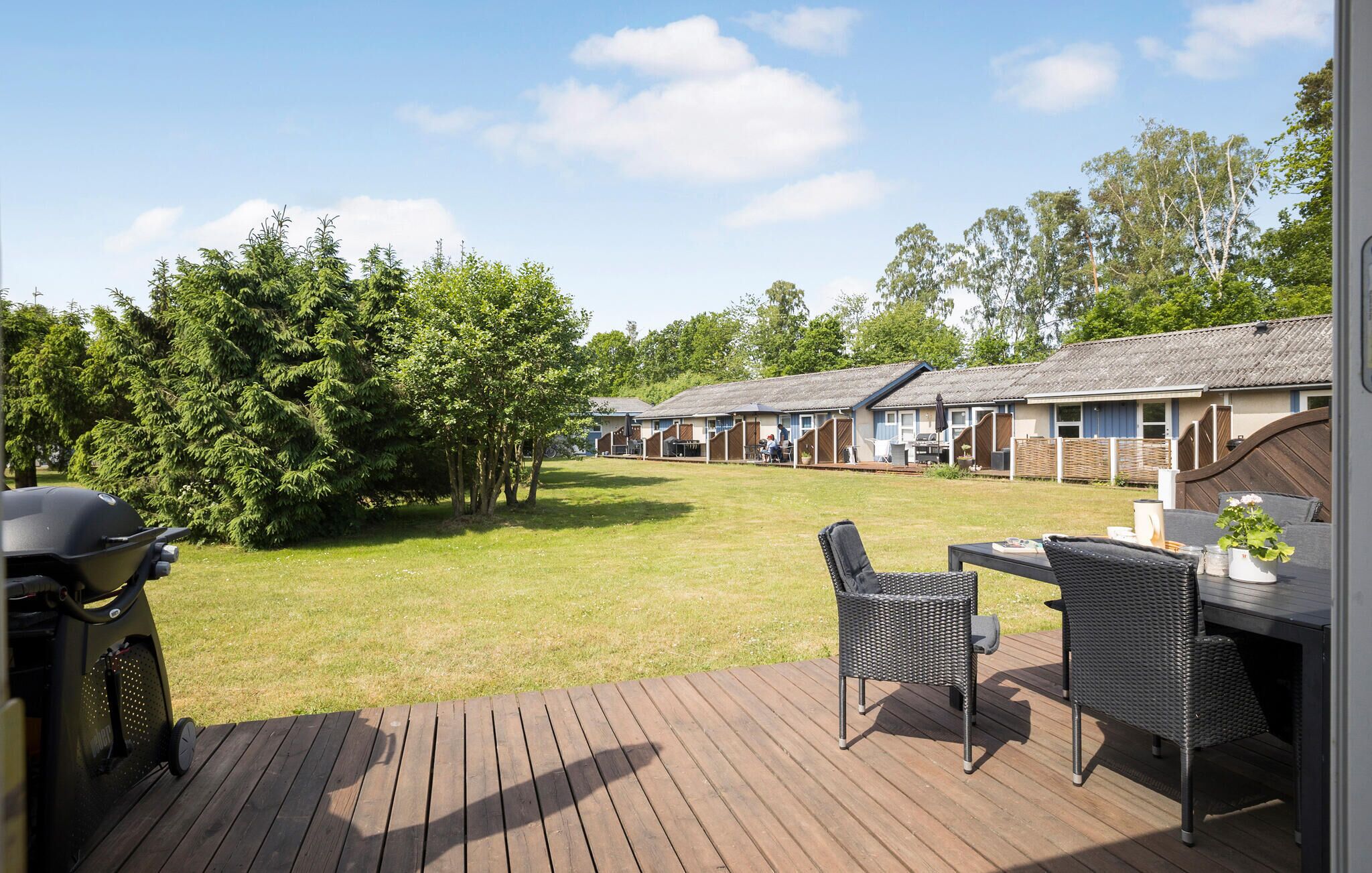 Eine Terrasse mit Gartenmöbeln, einem Grill und eine Reihe von Ferienhäusern mit Rasen und Bäumen im Hintergrund.