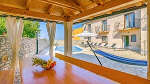 Una pérgola de madera con una mesa y una cesta de fruta, con vistas a una piscina y un edificio con balcones.
