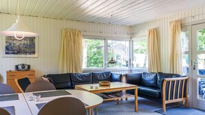 A cozy living room with a sofa, a dining table, and a coffee table. There is a window with curtains, a painting on the wall, and a door with a view of the outside.