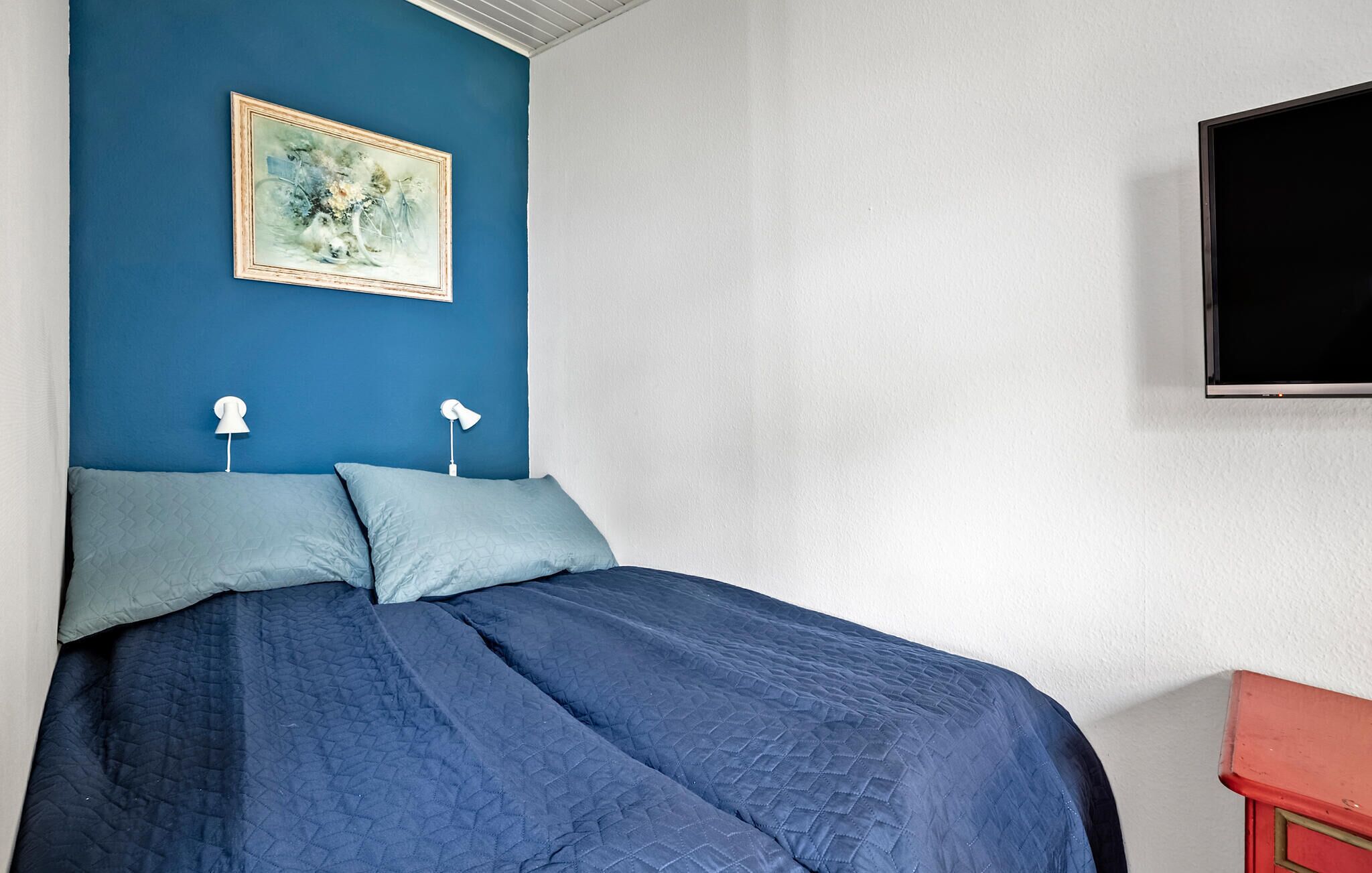 A bedroom with a bed, two bedside lamps, a painting on the wall, and a TV mounted on the wall.
