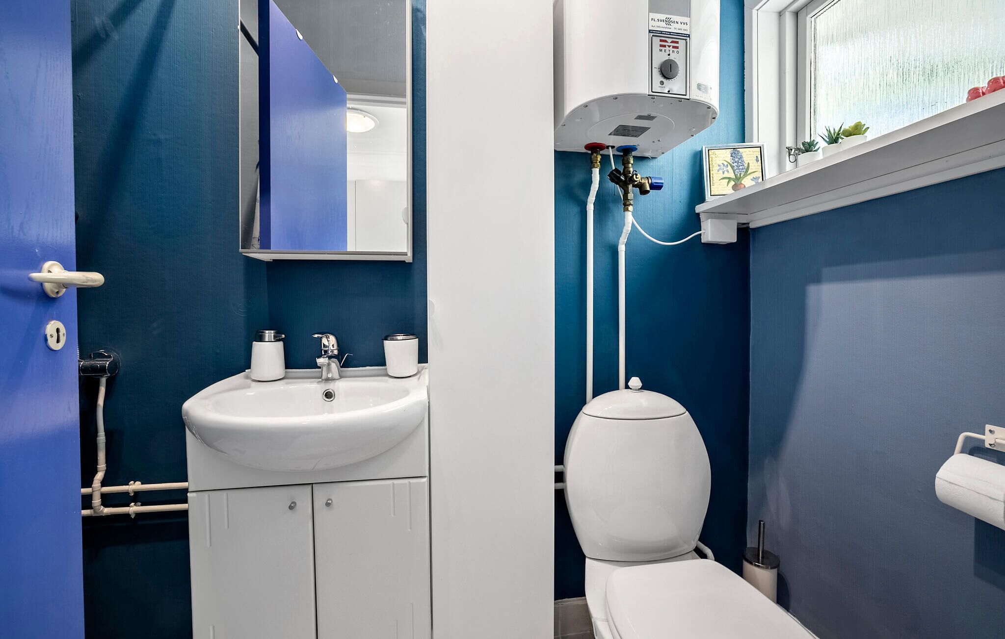 A bathroom with a white sink, toilet, and a mirror. There is a window above the toilet and a shelf with a picture frame.