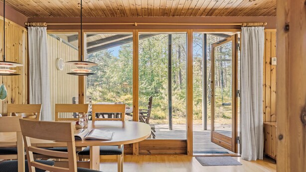 Un intérieur en bois comprenant un coin repas, de grandes portes-fenêtres coulissantes donnant sur une terrasse extérieure et une vue sur une zone boisée.