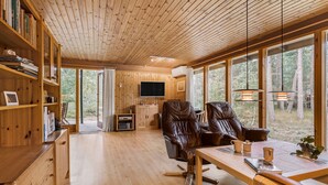 L’intérieur chaleureux d’un chalet avec des murs et des planchers en bois, une grande fenêtre donnant sur la forêt, un téléviseur et des fauteuils en cuir.