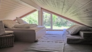 Une chambre mansardée chaleureuse avec un plafond en pente, une grande fenêtre donnant sur la verdure, un lit avec des oreillers et un panier en osier.