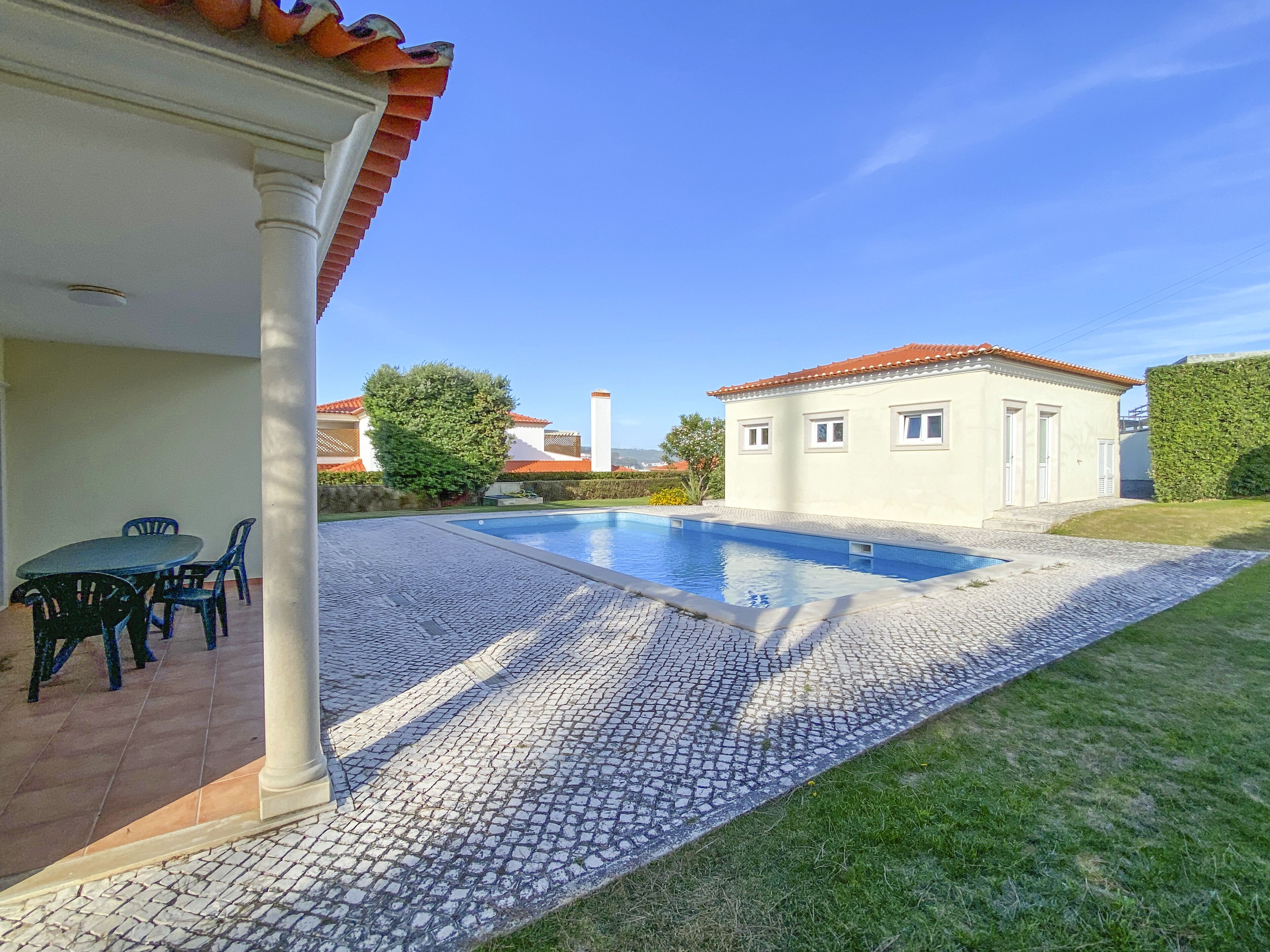 Une piscine avec une terrasse carrelée, un patio couvert avec des chaises et un bâtiment surmonté d’un toit recouvert de tuiles rouges.