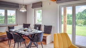 A dining area with a table set for four, surrounded by chairs, large windows, and a view of a garden.