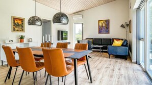 A modern living room with a black sofa, orange chairs, and a dining table with orange chairs. There is a painting on the wall and a window with a view of greenery. - Lovely home in Nexø with WiFi (Nexø)