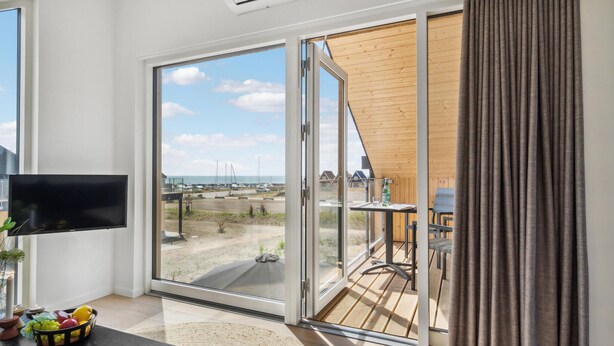 A modern room with a TV, a fruit bowl, and a view of a beach and boats through large glass doors.