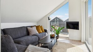 A modern living room with a grey sofa, a small table with a bowl of fruit, and a potted plant. A TV is mounted on the wall, and there is a large window providing a view of a building outside.