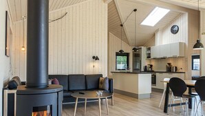 En hyggelig stue med pejs, sofa og sofabord. Der er et køkken med moderne hårde hvidevarer og et spisebord med stole.