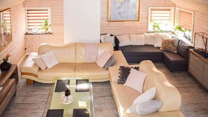 A cozy loft-style living room with a beige sofa, a dark sofa, and a glass coffee table. There are two windows with blinds, a painting on the wall, and a small plant on the windowsill. - Lovely home in Huskvarna (Huskvarna)