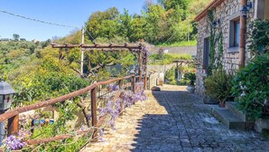 A cobblestone pathway leading to a stone building with a wooden pergola covered in purple flowers. - Stunning home in Ficarra (Ficarra)