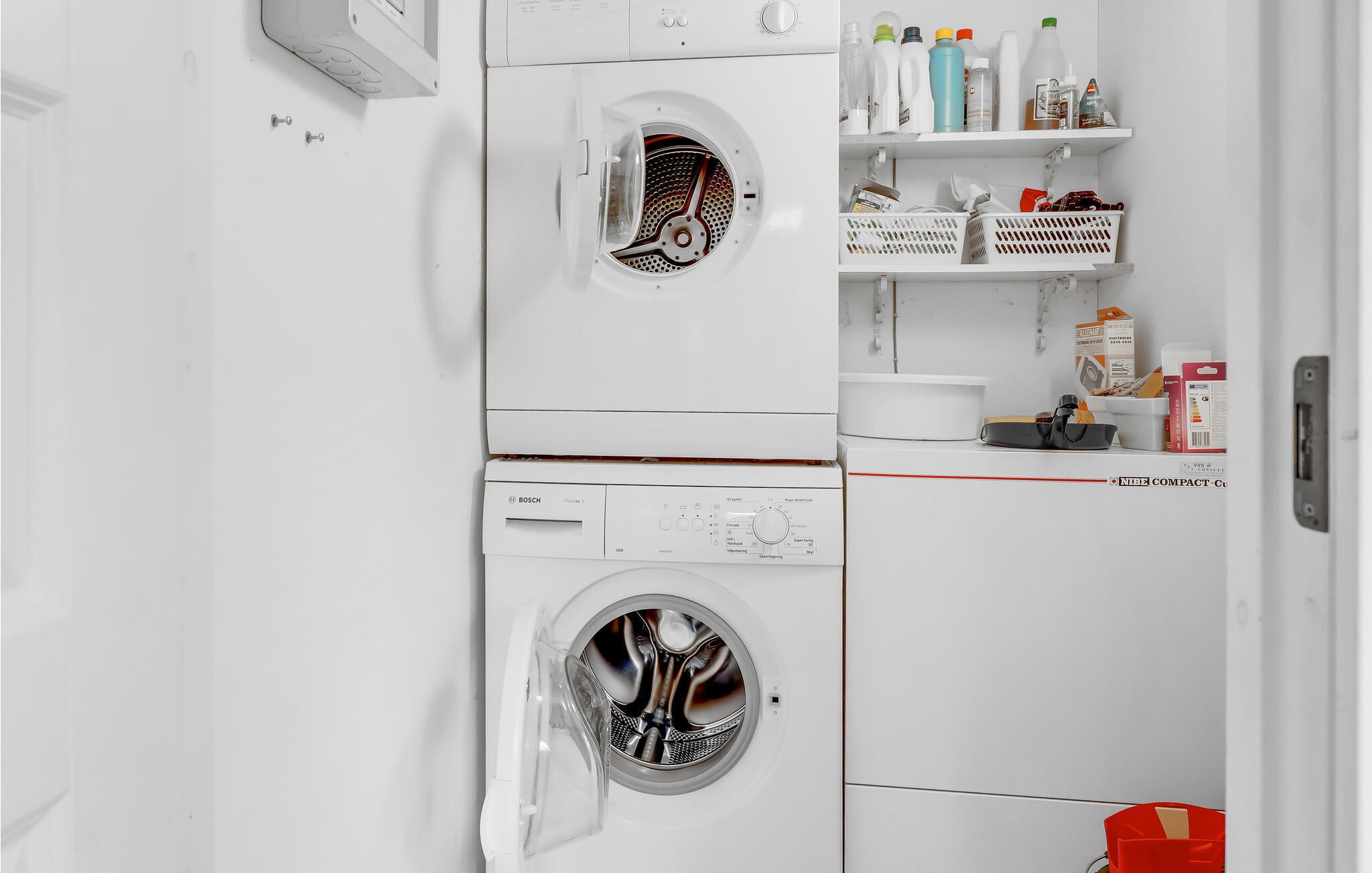 A compact, white laundry room with a Bosch washing machine, a dryer, and a shelf with cleaning supplies.
