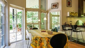Un coin repas avec une table recouverte d’une nappe jaune, entourée de chaises, et de grandes fenêtres donnant sur l’extérieur.