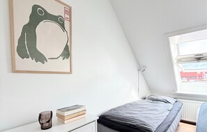 Un dormitorio con una cama, una mesita de noche con libros, un cuadro enmarcado en la pared y un ventanal con vistas a edificios.