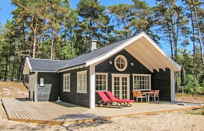 En stuga med en veranda, två fönster och en liten uteplats med stolar.