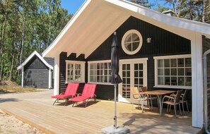 Ett trädäck med röda solstolar och ett bord, en täckt uteplats och en byggnad med stora fönster och ett runt fönster.