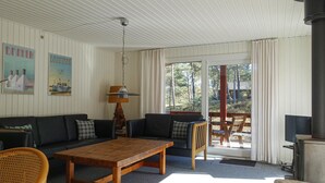 A cozy living room with a wooden table, two sofas, a wicker chair, and a large window with a view of trees and a building. - Pet friendly home in Nexø with kitchen (Nexø)