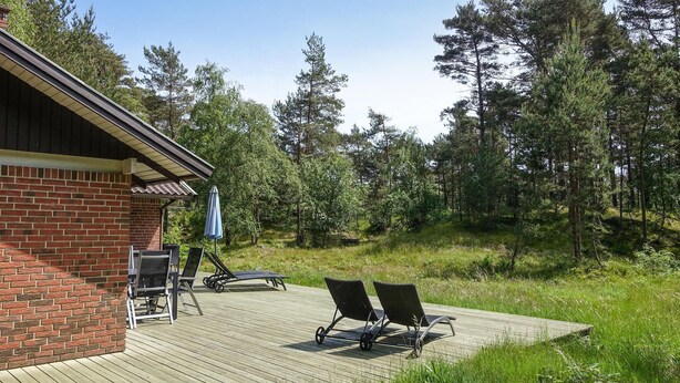 A wooden deck with two lounge chairs and a table, a brick building, and a forested area in the background. - Stunning home in Nexø with WiFi (Nexø)