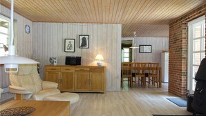 A cozy living room with a wooden ceiling, a sofa, a dining table, and a kitchen area with a brick wall and a refrigerator. - Stunning home in Nexø with WiFi (Nexø)
