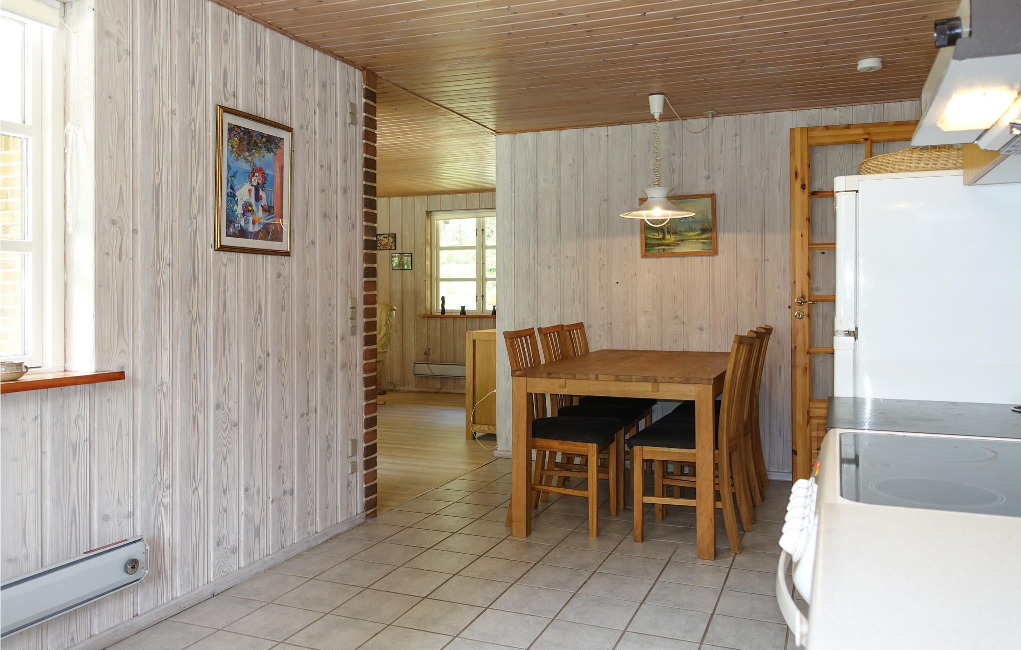 Uma sala de jantar com uma mesa e cadeiras de madeira, uma luminária pendente e uma geladeira.