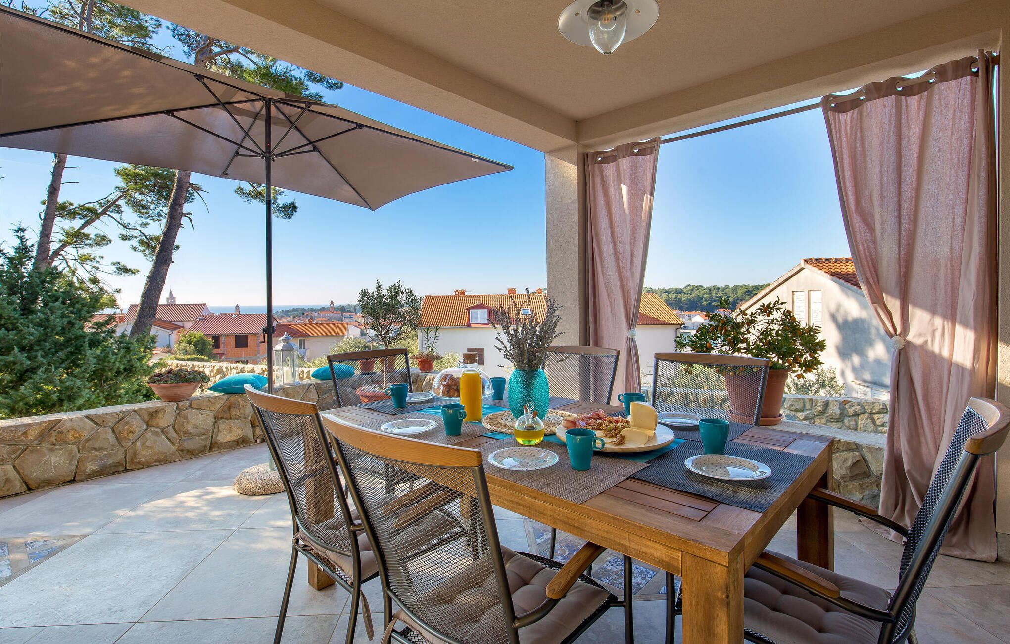 A patio with a table set for breakfast, outdoor chairs, a large umbrella, and a view of the surrounding area.