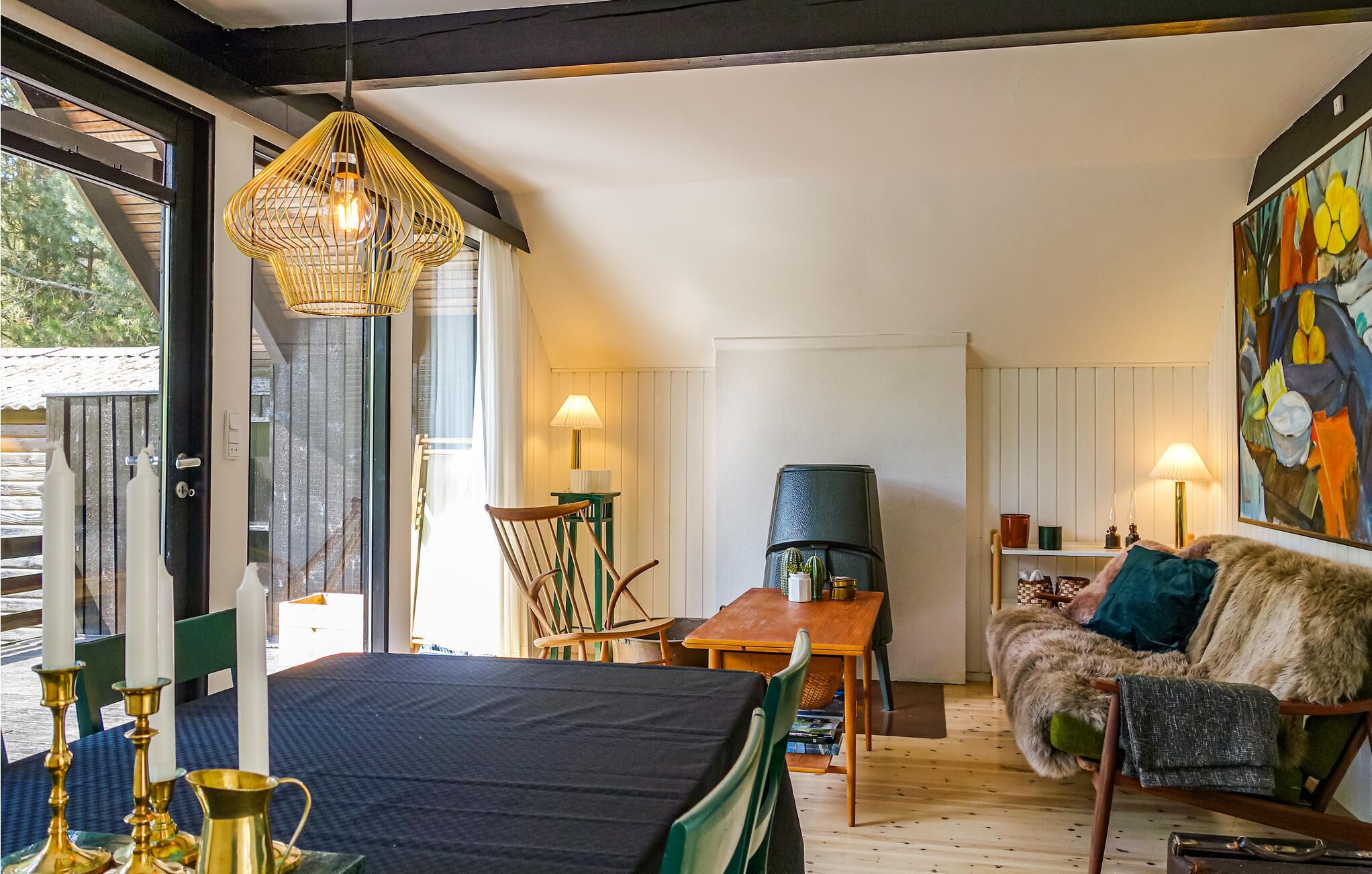 Ein gemütliches Wohnzimmer mit einer Couch, einem Tisch und einem Sessel. Ein großes Fenster und eine Tür führen nach drau ßen.
