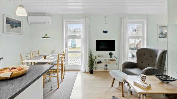 Uma sala de estar moderna com televisão, uma poltrona cinza, área de jantar com cadeiras de madeira e bancada da cozinha com ovos.