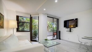 A modern living room with a white sofa, a glass coffee table, a TV mounted on the wall, and a balcony with potted plants. - KEY BISCAYNE LUXURY RETREAT - FULLY REMOLEDED 3 BEDROOM 2,000 SF RESIDENCE (Miami)