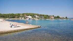 A coastal beach with clear waters, a sandy shore, and buildings in the background.