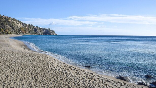 A beach with clear blue water, sandy shore, and a rocky cliff with buildings.