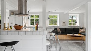 A modern kitchen with a central island, a dining area with a sofa and table, and large windows offering views of greenery. - Stunning home in Hasle with WiFi (Hasle)