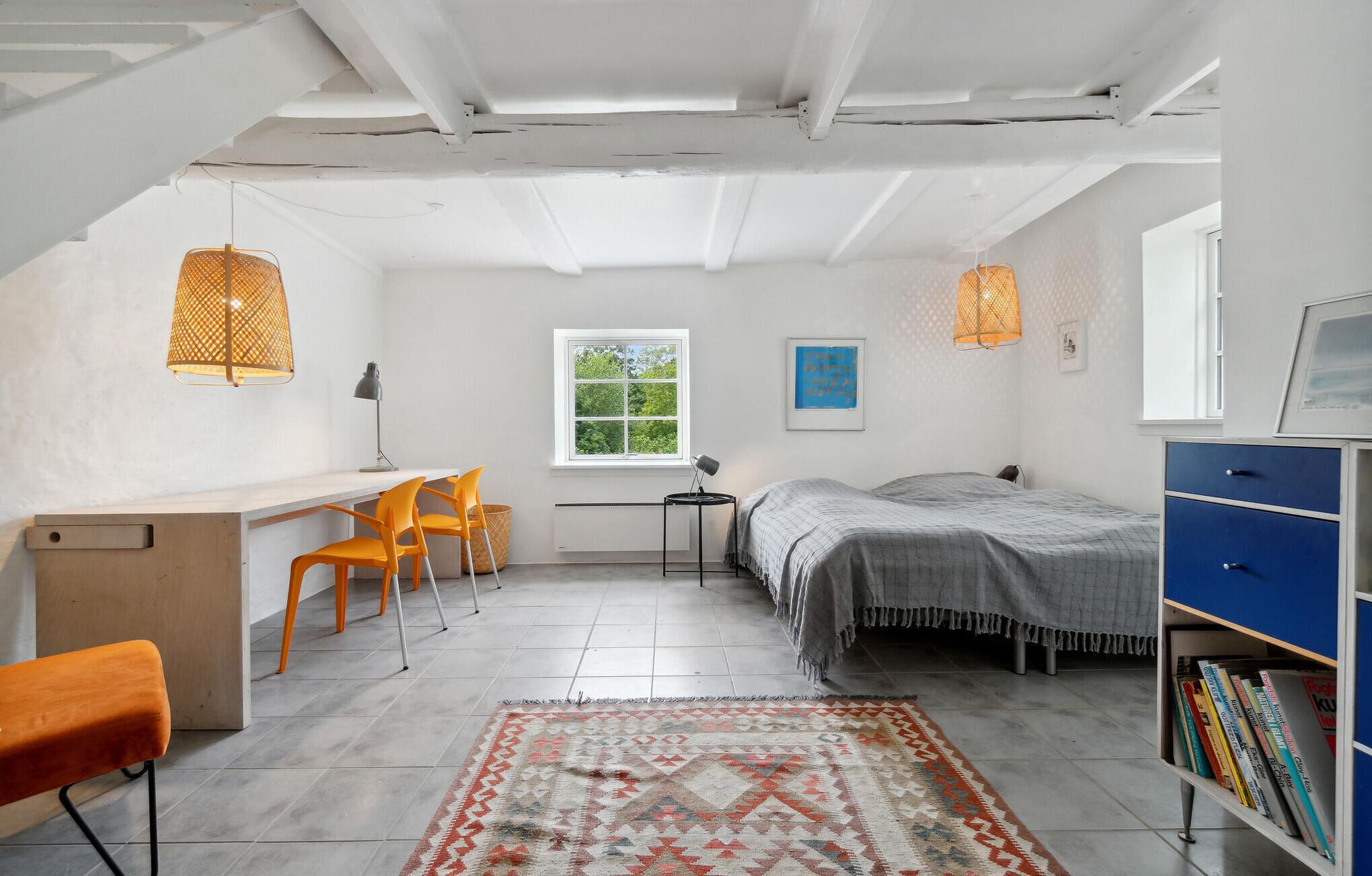 Une pièce compacte et bien éclairée, comprenant un bureau, deux chaises, un lit, une bibliothèque et une fenêtre laissant entrer la lumière naturelle.