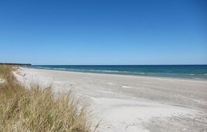 Ein Strand mit Sand und vereinzelten Grashalmen, ein klarer Himmel und ruhige Wellen.