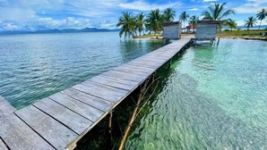 Eine hölzerne Pier erstreckt sich über klares türkisblaues Wasser und führt zu Bungalows auf Stelzen, im Hintergrund sind Palmen zu sehen.