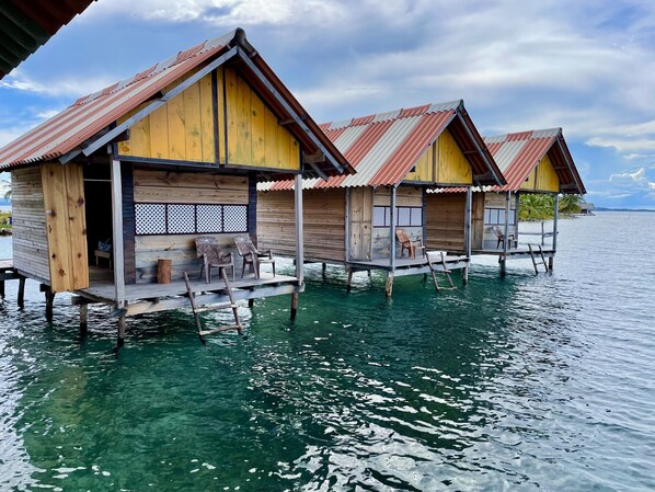 Cabañas sobre el agua con exteriores de madera y techos de metal, conectadas por pasarelas sobre aguas cristalinas.