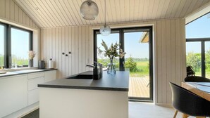 Une cuisine moderne dotée d’un grand îlot central, d’un coin repas avec une chaise noire, et offrant une vue sur un paysage verdoyant grâce à de grandes fenêtres.