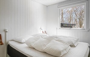 A bedroom with a bed, white walls, a window with a view of trees, and a small shelf with decorative items. - Amazing home in Aakirkeby (Aakirkeby)
