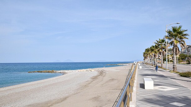 Une promenade en bord de mer, avec une plage de galets, des palmiers et un ciel bleu azur.