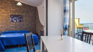 Une pièce avec un mur en pierre, un lit avec une literie bleue, une table à manger et une vue sur la mer par de grandes fenêtres.