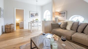 A spacious living area with a sofa, coffee table, and dining table. There is a wicker basket and a lamp on the table. - Witthüs, Whg. 19 - Witthüs (Wenningstedt-Braderup (Sylt))