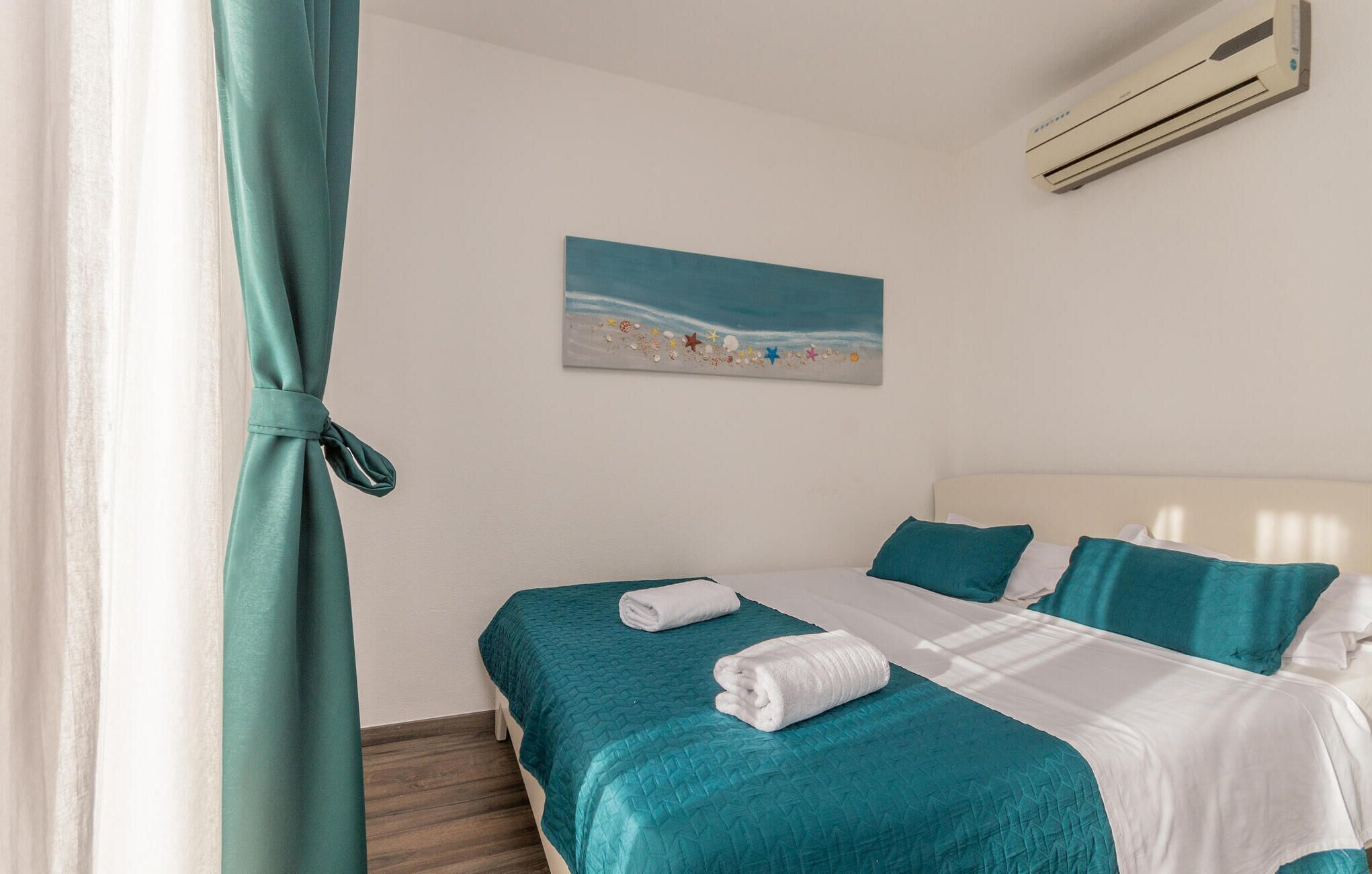 Camera d'hotel con un letto, biancheria da letto color petrolio, un quadro raffigurante una spiaggia e un condizionatore d'aria.