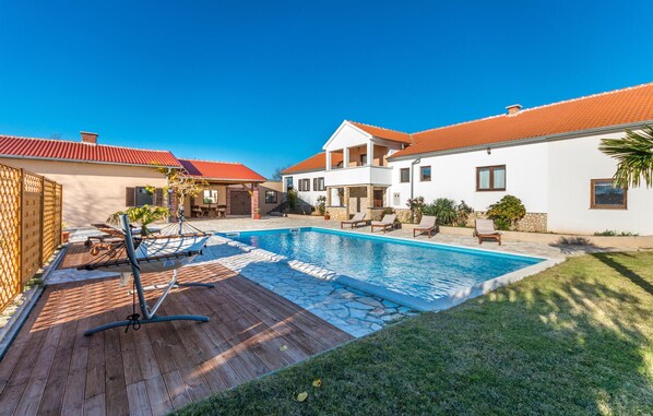 A swimming pool with lounge chairs, a wooden deck, and a stone pathway leading to a white house with a red roof.