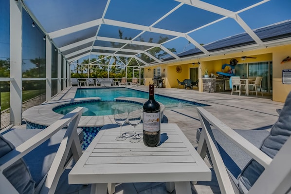 A pool area with a covered seating area, a bottle of wine, and a poolside table.