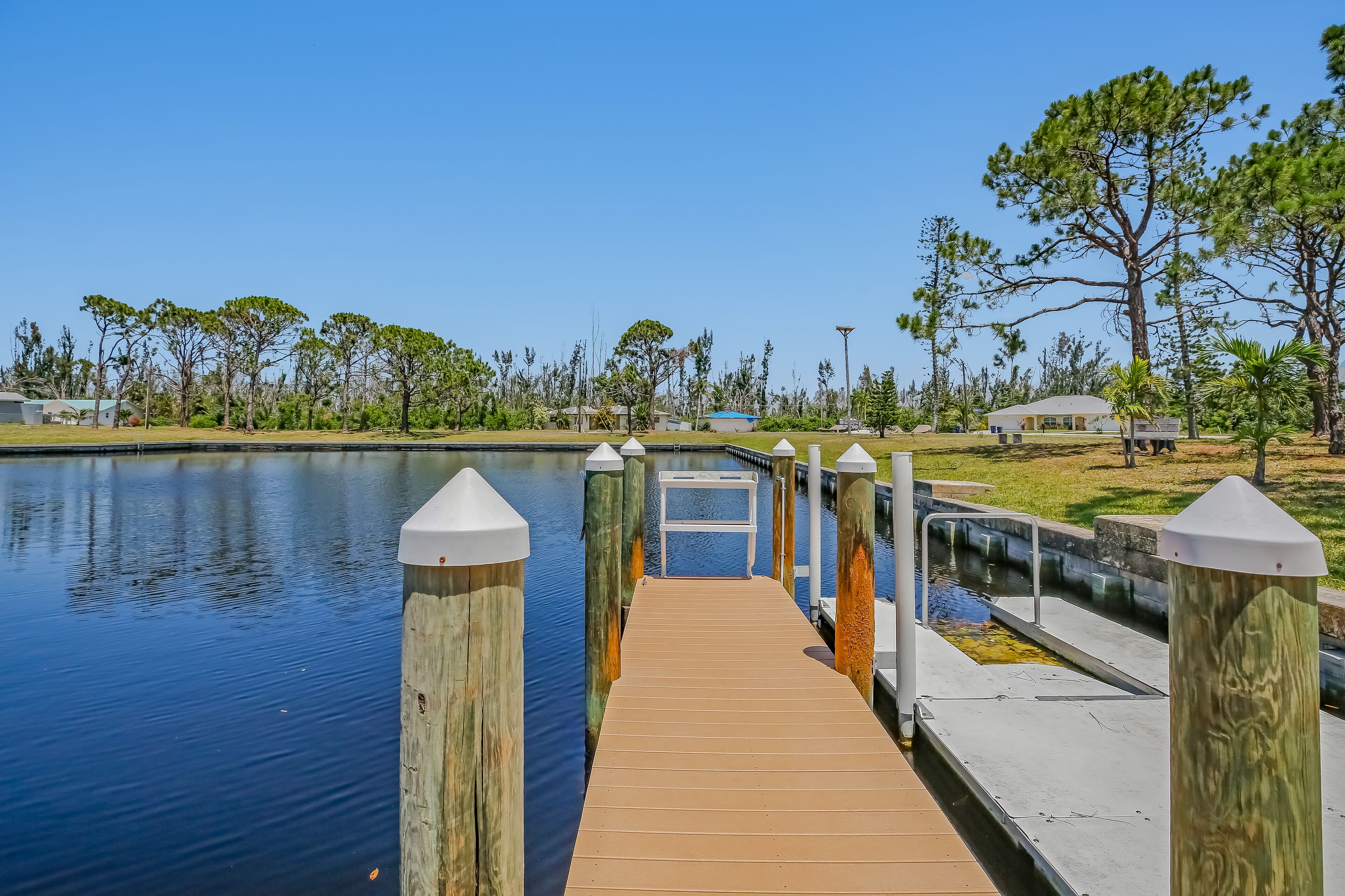 A wooden dock leading to a small white table by a calm body of water, surrounded by trees and a grassy area.