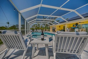 Uma área externa coberta com piscina, móveis brancos e uma garrafa de vinho sobre uma mesa.