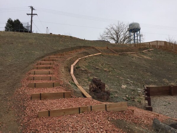 A winding pathway with wooden steps leading up a grassy hill, a water tower in the background, and a wooden fence along the top. - Mountain-View 5BR Retreat: Gym, Hot Tub, BBQ, Firepit, Playground, Sleeps 12 (Colorado Springs)