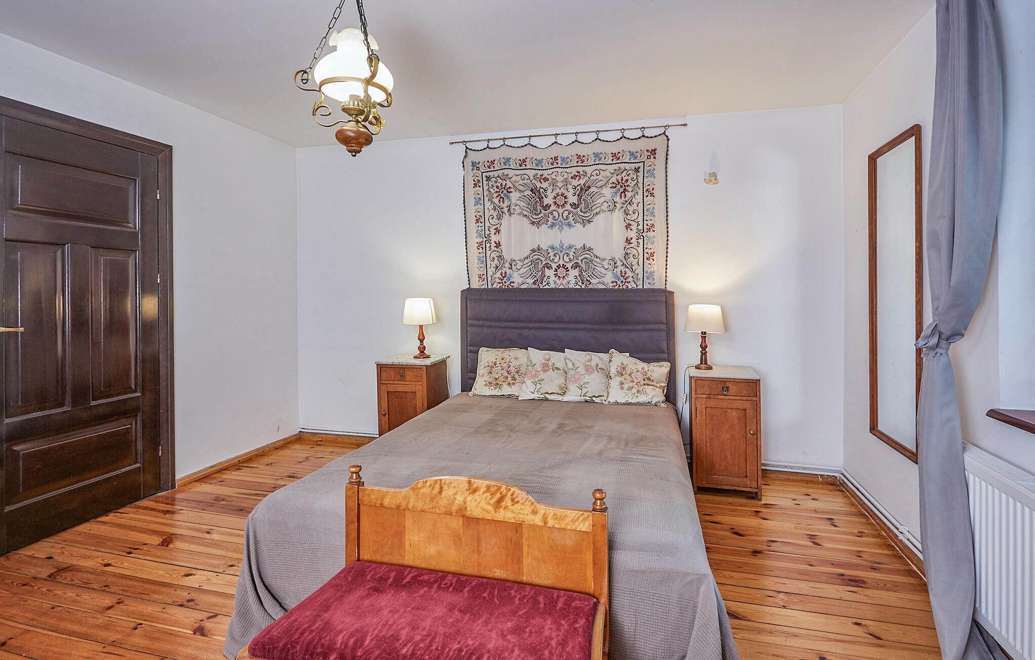 Una camera da letto con un letto di legno, due comodini, una lampada a sospensione, un arazzo appeso al muro e una porta di legno scuro.