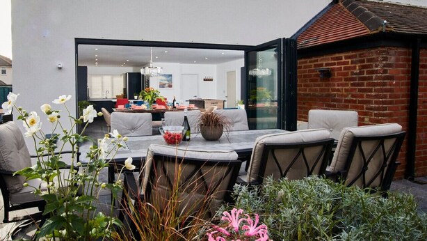 Een terras buiten met een eettafel en stoelen, een glazen constructie die naar een keuken leidt, en een bakstenen muur.
