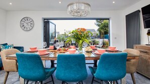 Een moderne eetruimte met een grote tafel, blauwgroene stoelen, een kroonluchter en uitzicht op de stad door grote ramen.
